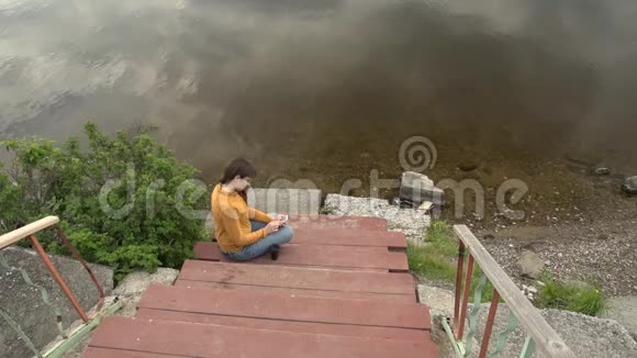 夏天一个年轻的女人坐在池塘边的码头上玩电话视频的预览图