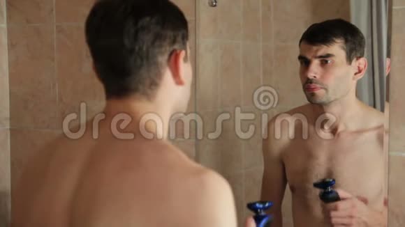 年轻人在浴室里用电动剃须刀刮胡子他在浴室里用剃刀刮胡子看着里面视频的预览图