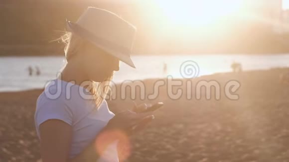戴着帽子的漂亮金发女孩在日落时在海滩上的手机上键入一条信息视频的预览图