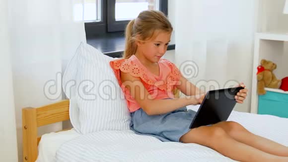 带着平板电脑的微笑女孩坐在家里的床上视频的预览图