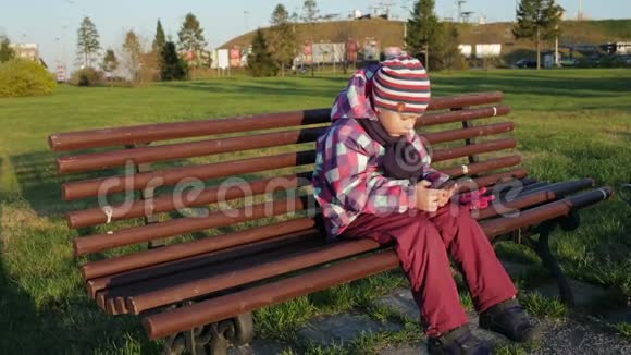 漂亮的小女孩在智能手机上使用应用程序坐在公园的长凳上视频的预览图
