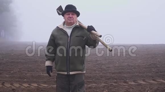 戴着帽子的高级农民手里拿着一把泥泞的锄头和田野上的草叉体力劳动福克视频的预览图