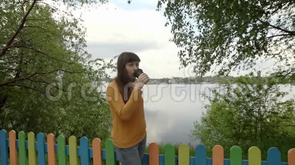 穿着黄色高领毛衣的年轻女子在池塘边喝咖啡视频的预览图