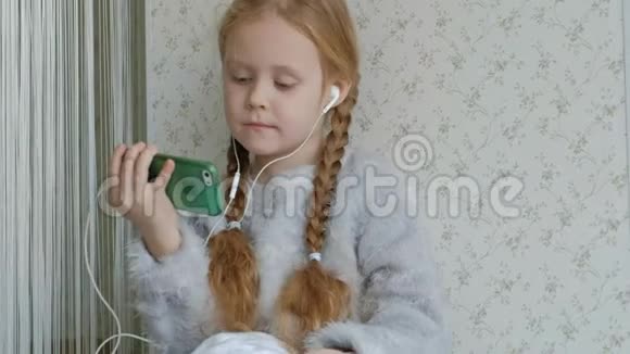 戴着耳机梳着辫子的快乐小女孩看着手机视频坐在窗台上的一个房间里视频的预览图