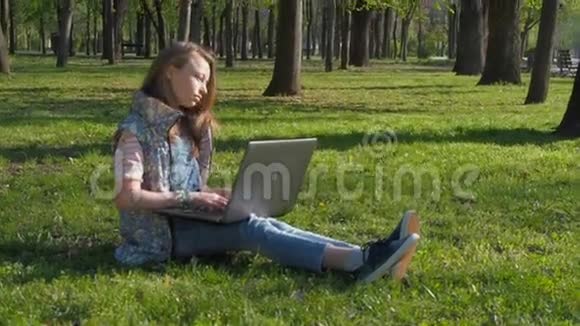 在大自然中做笔记本电脑的青少年那个女孩坐在公园的草地上在一个阳光明媚的日子里用笔记本电脑视频的预览图