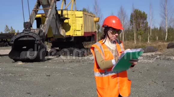 一名年轻的女工人站在一台挖掘机旁看着工程视频的预览图