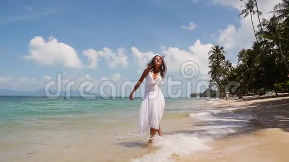 穿着白色连衣裙和花圈的漂亮年轻女人光着脚在海岸边奔跑享受着自己海滩视频的预览图
