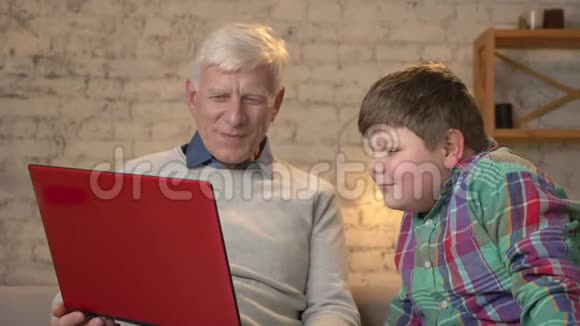 祖父和孙子坐在沙发上在笔记本电脑上看一部有趣的电影家庭舒适家庭懒散视频的预览图