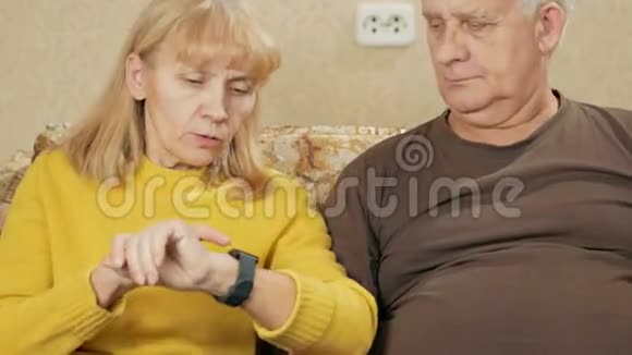年长夫妇在家里的沙发上看智能手表上的信息他们仔细阅读并阅读重要的文章视频的预览图