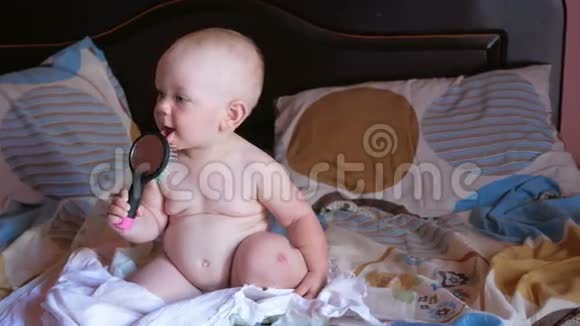 漂亮的宝宝在床上玩梳子微笑并试图梳理自己孩子一年梳子上有一面镜子视频的预览图