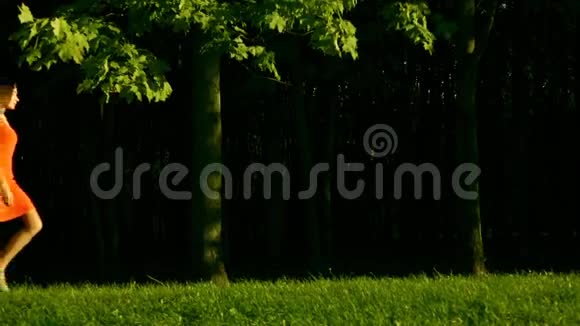 美丽的黑发女孩穿着红色连衣裙从左到右走在公园的草地上侧面看慢动作视频的预览图