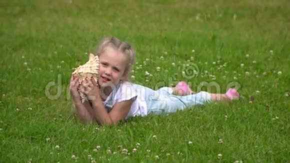 有一点金发女孩听着海浪在一个大公鸡壳美丽的小女孩躺在绿色的地上视频的预览图