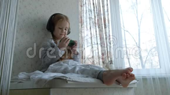 戴着耳机辫子的快乐小女孩看着手机视频坐在窗台上的一个房间里藏了一条毯子视频的预览图