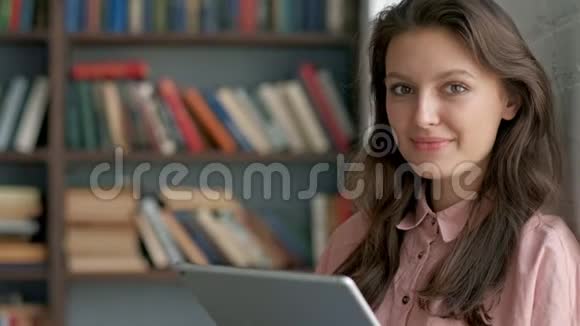 一位戴眼镜的卷曲年轻女子在图书馆对着相机微笑着使用平板电脑的特写镜头视频的预览图