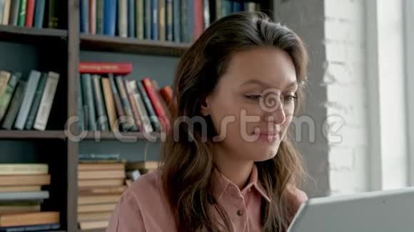 一位戴眼镜的卷曲年轻女子在图书馆对着相机微笑着使用平板电脑的特写镜头视频的预览图