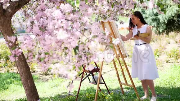 她手里拿着一幅美丽的女画家穿着白色的衣服画着一幅盛开的春苹果园里的鲜花视频的预览图