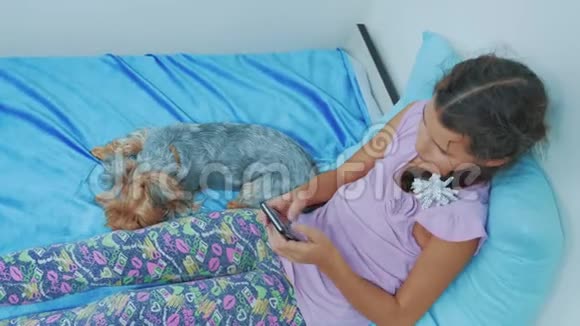 可爱的女孩和狗宠物躺在床上的智能手机肖像小女孩和狗宠物玩网络游戏视频的预览图