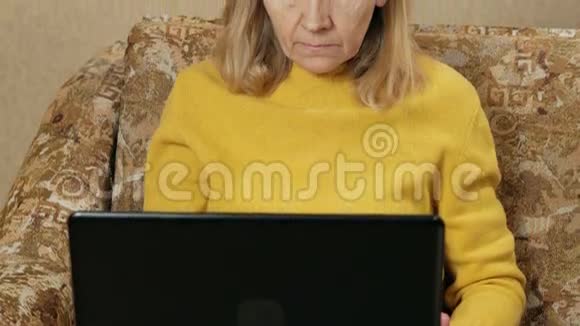 年老的女人在家里的沙发上小心翼翼地在笔记本电脑上工作她在键盘上打字看着屏幕这就是视频的预览图