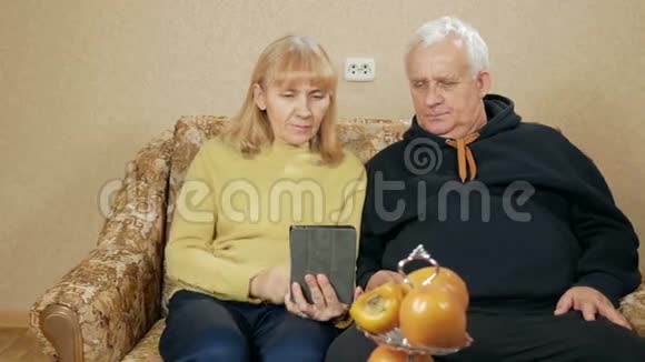 一对老夫妇手持数码平板电脑坐在客厅沙发上微笑的画像视频的预览图