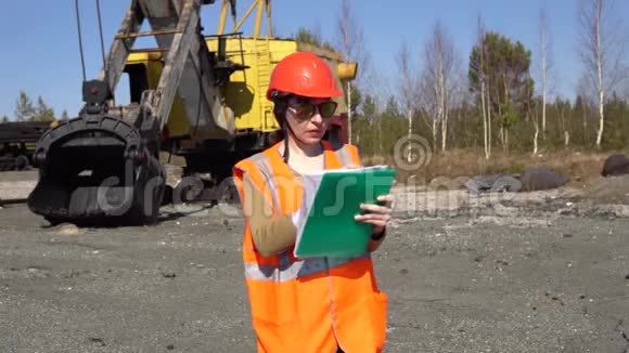 一名年轻的女工人站在一台挖掘机旁看着工程视频的预览图