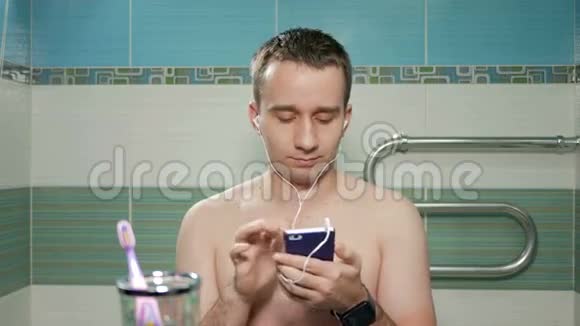 年轻迷人的男人在浴室里用耳机听音乐他唱着歌微笑着视频的预览图