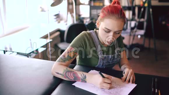年轻迷人的红头发妇女纹身艺术家坐在桌子上为室内工作室纹身创作素描视频的预览图