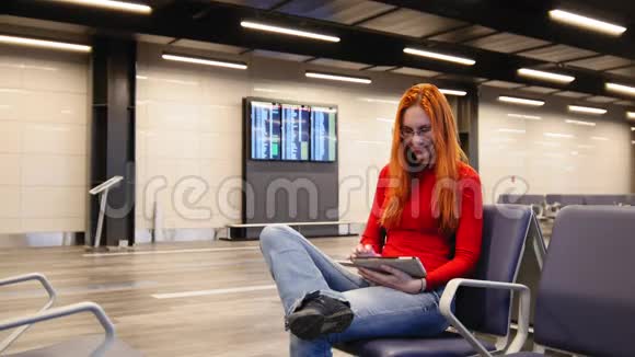 年轻漂亮的红头发和眼镜的女人在机场候机厅使用小玩意视频的预览图