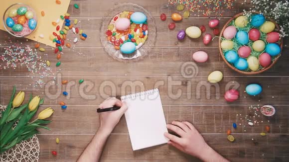 男人在桌上用复活节彩蛋装饰的笔记本上写复活节食谱顶部视图视频的预览图