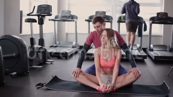 运动和有魅力的男人帮助一个漂亮的女人在健身房的地板上伸展视频的预览图