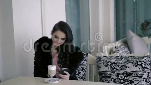 在露天咖啡馆里一个穿着皮草拿着拿铁和电话的聪明女人视频的预览图