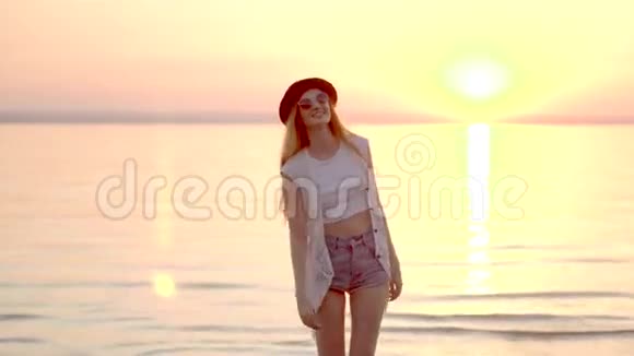 美丽的金发女孩戴着帽子在海洋和金色夕阳的背景上摆姿势视频的预览图