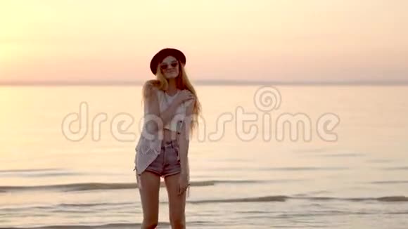 美丽的金发女孩戴着帽子在海洋和金色夕阳的背景上摆姿势视频的预览图