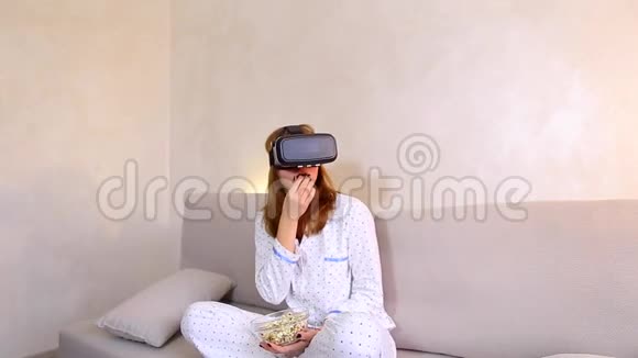 女孩看视频在VR眼镜与爆米花坐在沙发在明亮的卧室晚上视频的预览图
