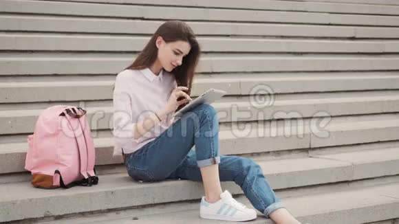年轻漂亮的时尚女孩坐在台阶上双手握在平板电脑上女生在社交网络中交流视频的预览图