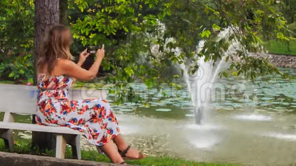 金发女孩坐在长凳上用Iphone拍喷泉视频的预览图