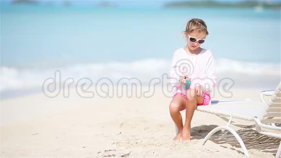 可爱的小女孩带着一瓶防晒霜坐在热带海滩上视频的预览图