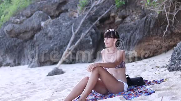 夏天的女孩年轻的女人穿着泳衣坐在靠近海洋和岩石的海滩上视频的预览图
