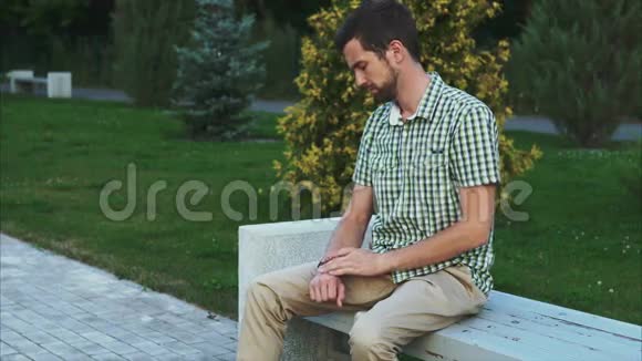 休闲打扮的人坐在公园的长凳上用他的现代智能手表视频的预览图