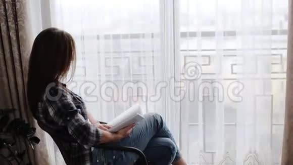 可爱的女人穿着蓝色牛仔裤和灰色T恤坐在窗前的桌子上在她的生活中看书视频的预览图