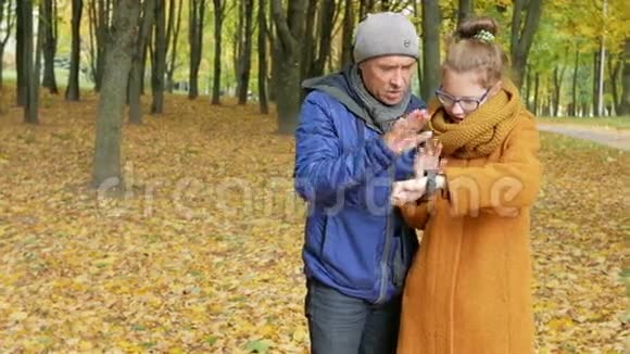 女儿少年教父亲在秋天公园里用智能手表工作爸爸仔细听重复一遍视频的预览图