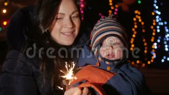 年轻的母亲带着一个孩子在街上用闪闪发光的东西庆祝新年拥抱并微笑着看着摄像机视频的预览图