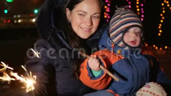 年轻的母亲带着一个孩子在街上用闪闪发光的东西庆祝新年拥抱并微笑着看着摄像机视频的预览图