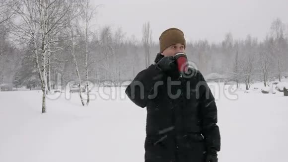 一个年轻人在冬天的森林里喝热水瓶里的热饮料大雪纷飞那个人看着镜头视频的预览图