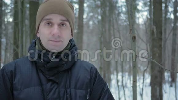年轻人看着镜头在冬天的森林里微笑冰雪景观一个穿深色夹克和暖帽的男人视频的预览图