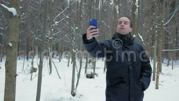 人在白雪皑皑的冬林里制作自拍手机他微笑着改变姿势以获得更好的照片可用性视频的预览图