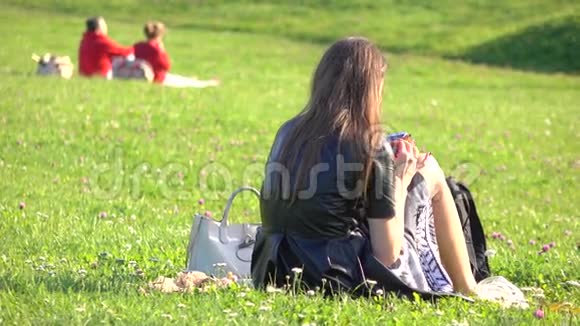 美丽的黑发女孩穿着黑色连衣裙在草坪上使用她的智能手机休闲时间4K视频视频的预览图
