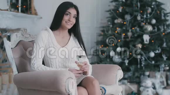 美丽的年轻女子坐在圣诞树旁边的椅子上香槟酒视频的预览图
