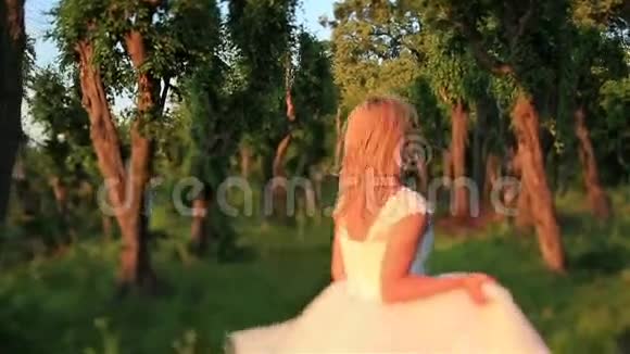 美丽的金发新娘在阳光照耀的绿色森林里奔跑美丽的日落在乡村背面视频的预览图
