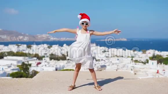 戴着圣诞老人红帽子的小女孩是迈科诺斯的户外背景典型的希腊传统村落街上的孩子视频的预览图