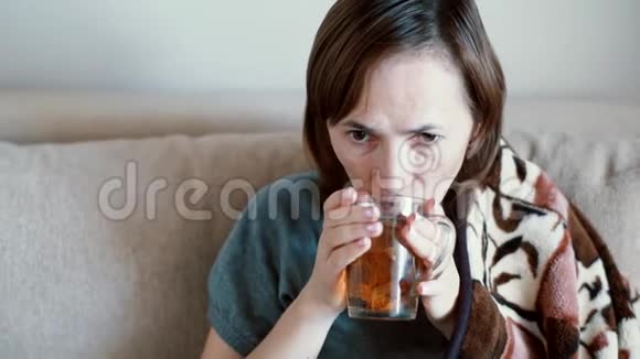 生病的女人在家里坐在沙发上盖着毯子喝着温暖的饮料视频的预览图